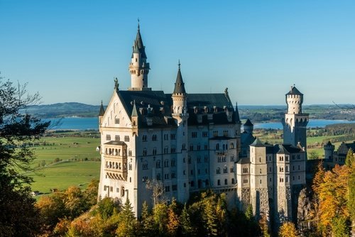 Schloss Neuschwanstein mit weißen Türmen über herbstlichem Wald, dahinter Seen und flaches Land